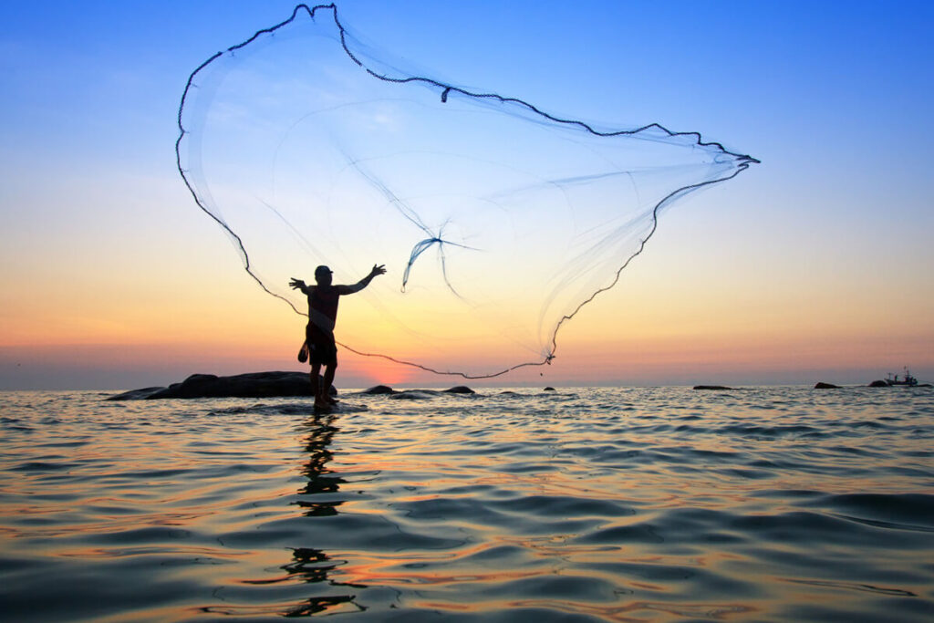 man throwing net to catch fish in the ocean with a beautiful sunset